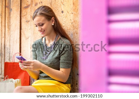 Close up portrait of an attractive young woman sitting by an old textured wooden door with shopping bags, joyfully smiling using a smartphone to network on vacation. Outdoors technology and lifestyle.