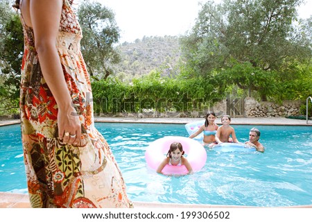 Family enjoying a summer holiday in a vacation home together, having fun in a swimming pool with dad while the figure of the mother walks through the foreground. Active family lifestyle outdoors.