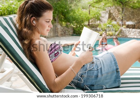 Close up side portrait view of a teenager girl relaxing and lounging during a summer holiday in a sunbathing lounger while reading a book and listening to music with headphones. Vacation lifestyle.