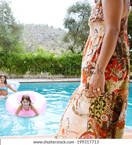Family enjoying a summer holiday in a vacation home together, having fun in a swimming pool with dad while the figure of the mother walks through the foreground. Active family lifestyle outdoors.