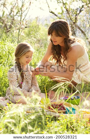 Mother and daughter enjoying a picnic together in a green field with flowers, eating strawberries with mom crouching to help the young girl. Family activities and healthy eating lifestyle, outdoors.