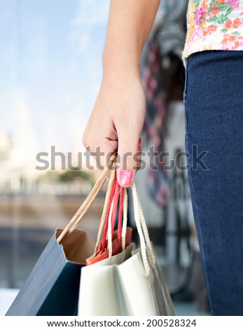 Close up detail view of a young woman middle section and hand holding paper carrier bags in a shopping mall with glass windows and city reflections during a sunny day. Consumer outdoors lifestyle.