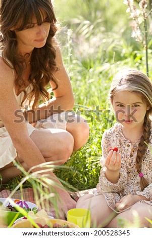 Close up of an attractive mother and daughter enjoying a picnic together in a green field, eating strawberries with mom crouching next to her child. Family activities and healthy lifestyle, outdoors.