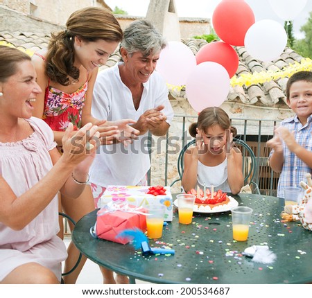 Family group enjoying and celebrating a young girl child birthday during a summer day, sitting around a table with a birthday cake and gifts and a surprised birthday girl. Family celebrating outdoors.