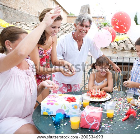 Family group enjoying and celebrating a young girl birthday during a summer day, sitting around a table with a strawberry birthday cake and gifts and throwing confetti. Family celebrating outdoors.