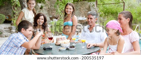 Panoramic view of a family group sitting together around a table with food and drink in a summer holiday garden swimming pool, posing for photos during a vacation together. Active family lifestyle.