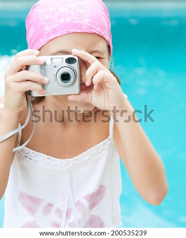 Close up portrait of a young child using a digital photo camera to take pictures pointing at the camera with a blue water swimming pool space in the background. Active children learning, outdoors.