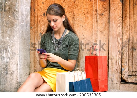 Close up portrait of an attractive woman sitting on an old aged building wooden door with her shopping bags, being thoughtful and using a smartphone to go on line, outdoors. Technology and lifestyle.