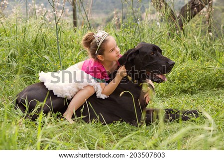 Profile view of a pretty young girl wearing a pink fancy dress, sitting on her dogs back enjoying a sunny holiday in a green park field, hugging him outdoors. Active family with pets, lifestyle.