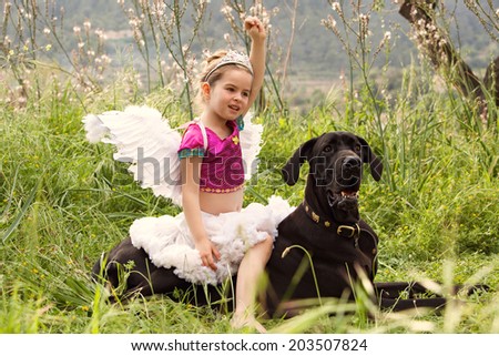 Young child girl dressing up in a fancy dress with wings, sitting on her great dane dogs back holding her fist up like a heroine. Proud owner playing with her pet enjoying a summer holiday together.