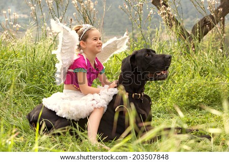 Side view of a young girl wearing a pink fancy dress with wings, sitting on her dogs back enjoying a sunny holiday in a green park field, smiling outdoors. Active family with animal pets, lifestyle.