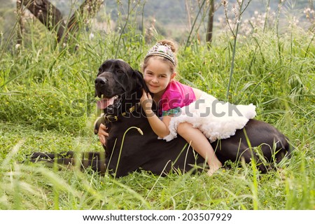 Profile view of a young girl wearing a pink fancy dress with crown, sitting on her dogs back enjoying a sunny holiday in a green park field, hugging and smiling. Active family with pets, lifestyle.