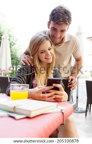Portrait of an attractive young couple on holiday sitting at a cafe terrace table using a smartphone and smiling together, visiting a destination city on a summer vacation. Technology lifestyle.