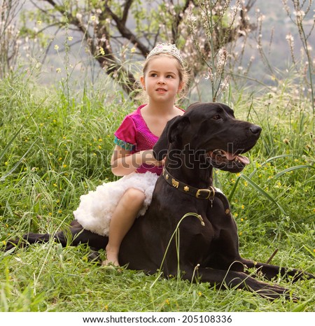 Young child girl dressing up in fancy dress in a green nature field, sitting on her great dane dogs back being thoughtful. Happy dog owner playing with her pet and enjoying a summer holiday together.