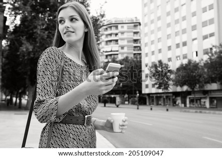 Black and white portrait of an attractive young professional business woman standing in a city street holding and using a smartphone on her way to work, turning back. Business technology on the go.