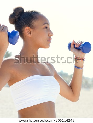 Side portrait of a fit healthy beautiful teenager girl exercising on a white sand beach, lifting small weights during an exercise and sport routine on a sunny day. Sporty healthy lifestyle outdoors.