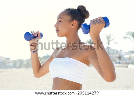 Side portrait of a fit healthy beautiful teenager girl exercising on a white sand beach, lifting small weights during an exercise and sport routine on a sunny day. Sporty healthy lifestyle outdoors.
