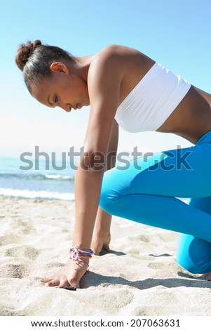Close up side view of a healthy african american woman crouching in the ready position to start running exercise training against a blue sea and sky during a sunny day. Sport lifestyle outdoors.