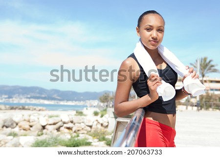 Portrait of an athletic african american young woman relaxing with a towel, leaning on a railing exercising by the seaside during a sunny summer day. Health and sport lifestyle and wellness outdoors.