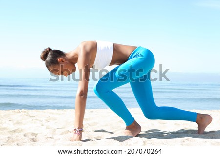 Side view of attractive healthy young african american woman crouching in the ready position to start running exercise training against a blue sea and sky during a sunny day. Sport lifestyle outdoors.