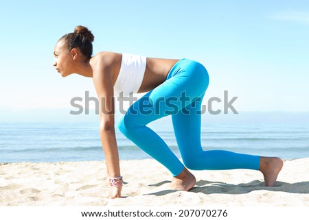Side view of attractive healthy young african american woman crouching in the ready position to start running exercise training against a blue sea and sky during a sunny day. Sport lifestyle outdoors.