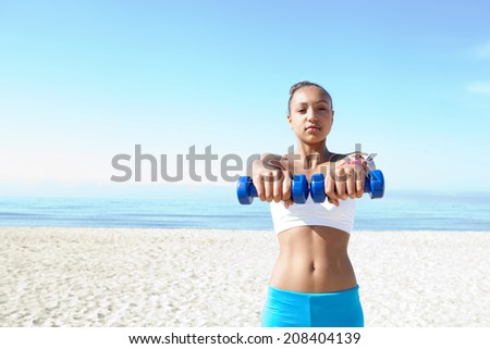 Portrait of a healthy beautiful teenager girl exercising on a white sand beach, lifting weights during an exercise and sport routine against an intense blue sky. Sporty healthy lifestyle outdoors.