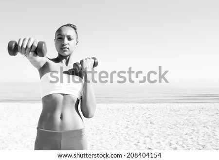 Black and white portrait of a beautiful teenager girl exercising on a sandy beach, lifting small weights during an exercise and sport routine against a sunny sky. Sporty healthy lifestyle outdoors.