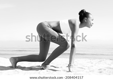 Black and white view of a young african american woman crouching in the ready position to start running exercise training against a blue sea and sky during a sunny day. Sport lifestyle outdoors.