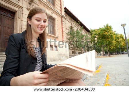Portrait of a professional business woman sitting at the entrance of an official office building reading a financial newspaper and smiling, outdoors. Corporate business in the financial city.