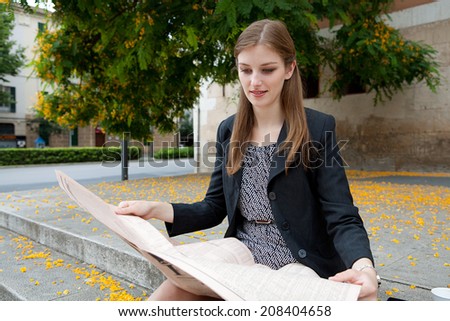 Portrait of a professional business woman sitting on the stone steps of an office building reading the stock market pages of a financial newspaper, smiling outdoors. Corporate business financial city.