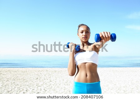 Portrait of a fit beautiful teenager girl exercising on a white sand beach, lifting small weights during an exercise and sport routine against an intense blue sky. Sporty healthy lifestyle outdoors.