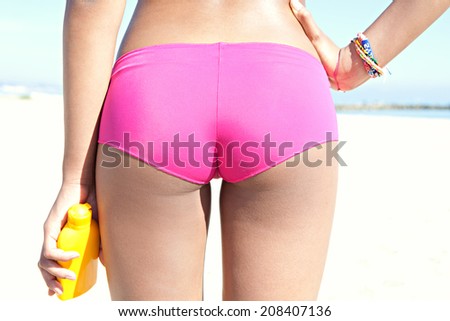 Sexy close up middle view of an attractive tanned young woman on a sandy beach, holding a bottle of sun protection body cream against a sunny blue sky on holiday. Colorful beauty lifestyle.