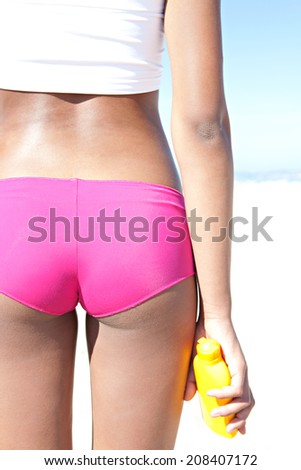 Sexy close up middle view of an attractive tanned young woman on a white sand beach, holding a bottle of sun protection body cream against a sunny blue sky on holiday. Beauty lifestyle.