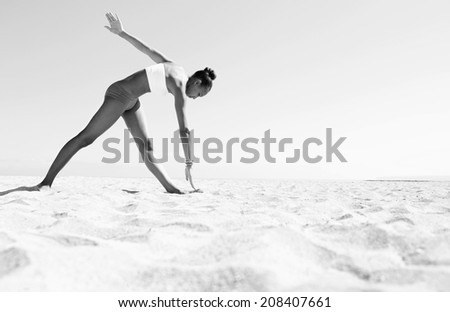 Black and white wide view of an athletic african american young woman exercising stretching her arms and legs against a blue sky on a sunny beach. Sport and healthy lifestyle, outdoors.