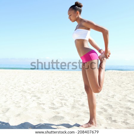 Side view of a healthy sporty young african american woman stretching her legs, exercising and training against a blue sea and sky during a sunny day on a sandy beach. Sport lifestyle outdoors.