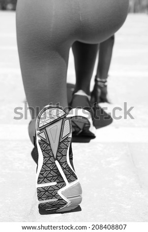 Close up black and white rear view of a young woman crouching down and preparing to run in the go position, exercising during a sunny day. Active sport woman jogging outdoors, lifestyle.