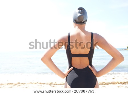 Rear faceless view of a young attractive woman back standing on a beach facing the sea, wearing swim wear and getting ready for swimming training, outdoors. Healthy sport and lifestyle.