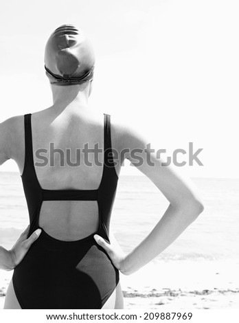 Rear black and white view of a young attractive woman back standing on a beach facing the sea, wearing swim wear and getting ready for swimming training, outdoors. Healthy sport and lifestyle.