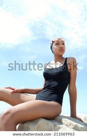 Side view of a fit, healthy young black woman swimmer sitting on a rock by the sea against a sunny blue sky, wearing swimming goggles and costume. Sport and training lifestyle, outdoors.