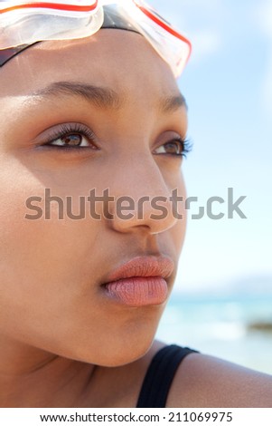 Close up portrait view of an attractive healthy young black woman swimmer sitting by the sea against a sunny blue sky, wearing swimming goggles and looking thoughtful. Sport lifestyle, outdoors.