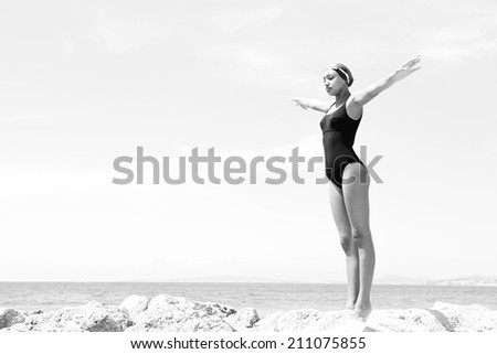 Black and white body view of a beautiful young woman swimmer diver preparing to dive off a rocky cliff by the sea, raising her arms up against a sunny blue sky. Health and beauty, sport lifestyle.