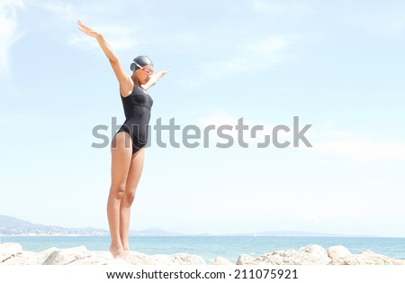 Side body view of a beautiful young woman swimmer diver preparing to dive off a rocky cliff by the sea, raising her arms up against a sunny blue sky. Health and beauty, sport lifestyle.