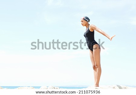 Profile body view of a beautiful young woman swimmer diver preparing to dive off a rocky cliff by the sea, wearing goggles against a sunny blue sky. Health and beauty, sport lifestyle.