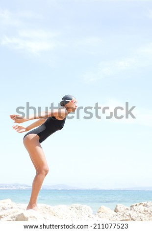 Profile body view of a beautiful young woman swimmer diver preparing to dive off a rocky cliff by the sea, wearing goggles against a sunny blue sky. Health and beauty, sport lifestyle.