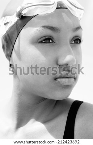 Close up black and white portrait view of an attractive healthy black woman swimmer sitting by the sea against a sunny sky, wearing swimming goggles and looking thoughtful. Sport lifestyle, outdoors.