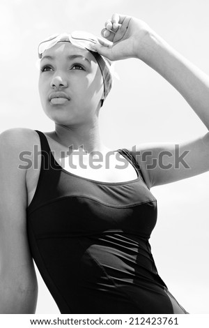 Black and white portrait view of a fit, healthy young black woman swimmer sitting by the sea against a sunny sky, holding swimming goggles and looking thoughtful. Sport lifestyle, outdoors.