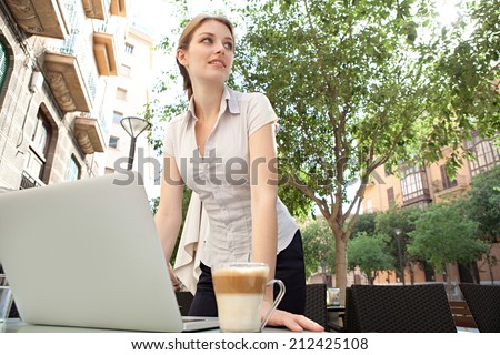 Beautiful professional young business woman standing at a coffee shop table, drinking a beverage and using laptop computer working in the financial city, outdoors. Business people at work.
