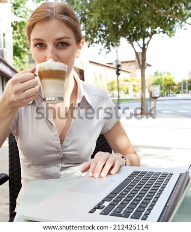 Portrait of an attractive young professional business woman sitting at a cafe terrace drinking  a hot beverage coffee and using a laptop computer in the financial district of a city, outdoors.