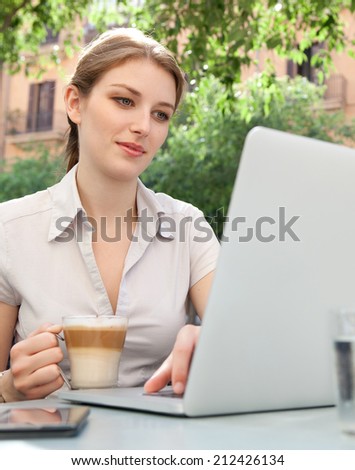 Attractive professional young business woman sitting at a coffee shop smiling, drinking a beverage and using laptop computer and smartphone technology in the financial city, outdoors.