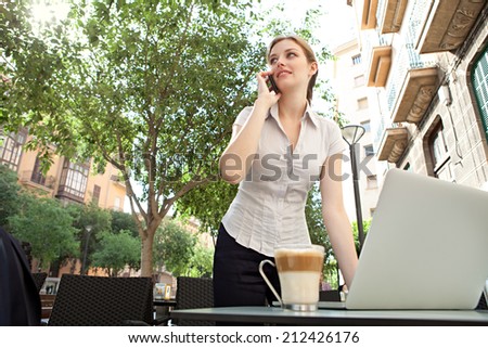 Beautiful professional young business woman standing at a coffee shop table, drinking a beverage, using laptop computer and making a phone call, working in the financial city, outdoors.